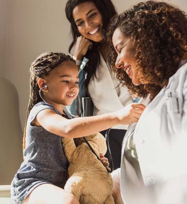 A pediatric patient listens to a physician's heartbeat through a stethoscope while her mother watches with a smile on her face.