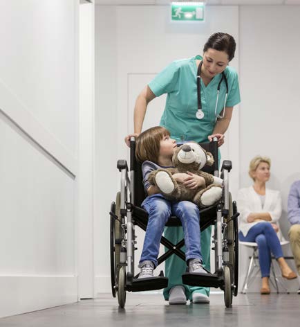 A clinician pushing the wheelchair of a pediatric patient holding a teddy bear.