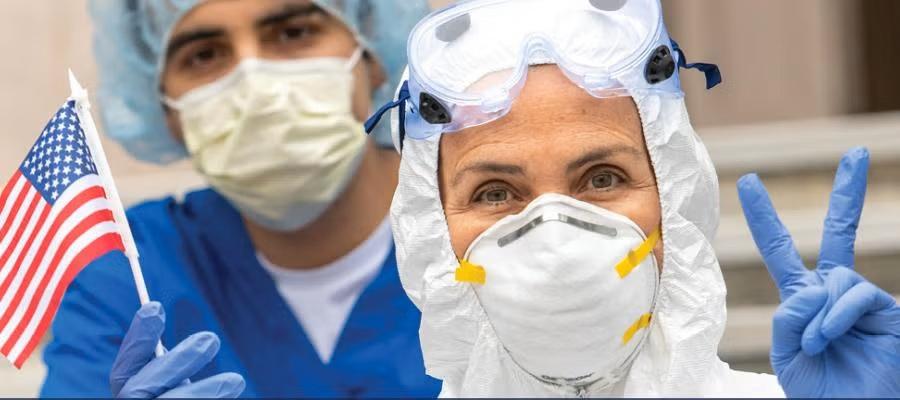 National Hospital Week. Two clinicians in surgical scrubs, masks, hoods, and gloves, one holding an American flag, one giving the peace sign.