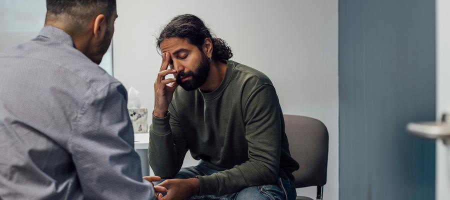 A man sits in a chair, looking distressed, while talking to a health care professional.